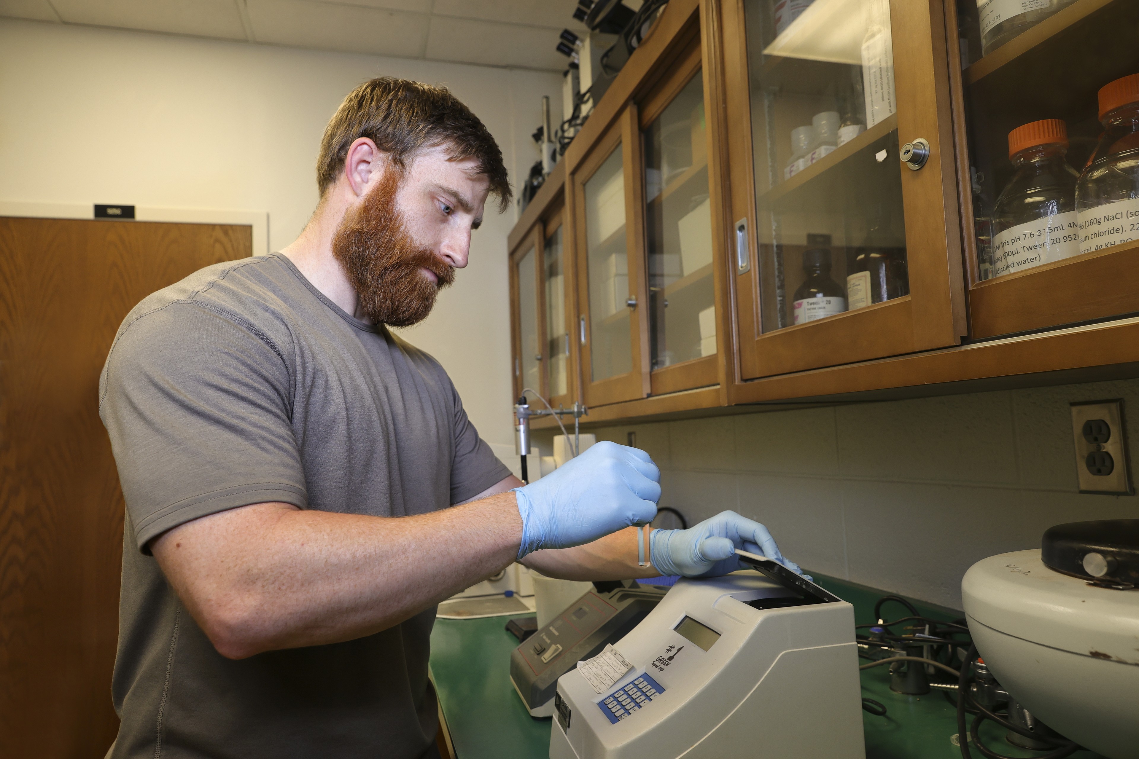 student, ryan wagner, wearing a grey tshirts, blue gloves testing a sample with equipment