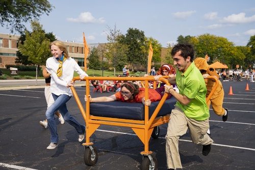 Students in costume push a rolling bed frame across a parking lot