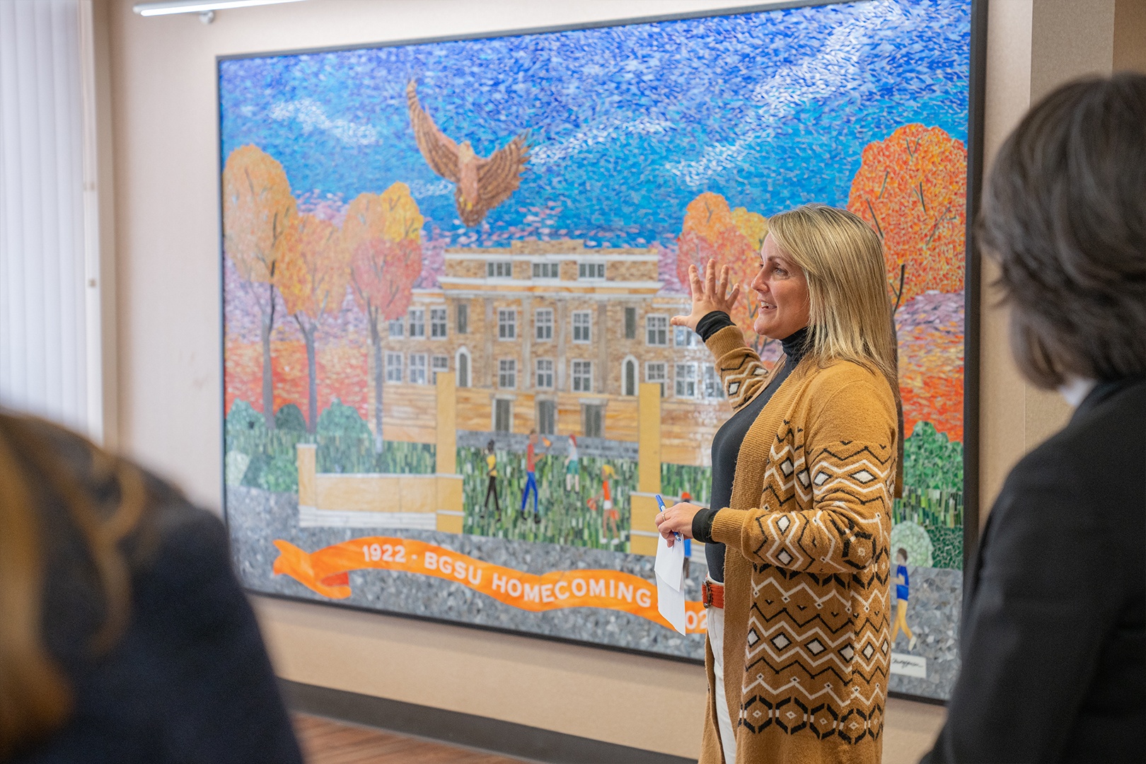 BGSU alumna Sarah Brokamp stands in front of a mosaic artwork depicting University Hall, students, and a falcon
