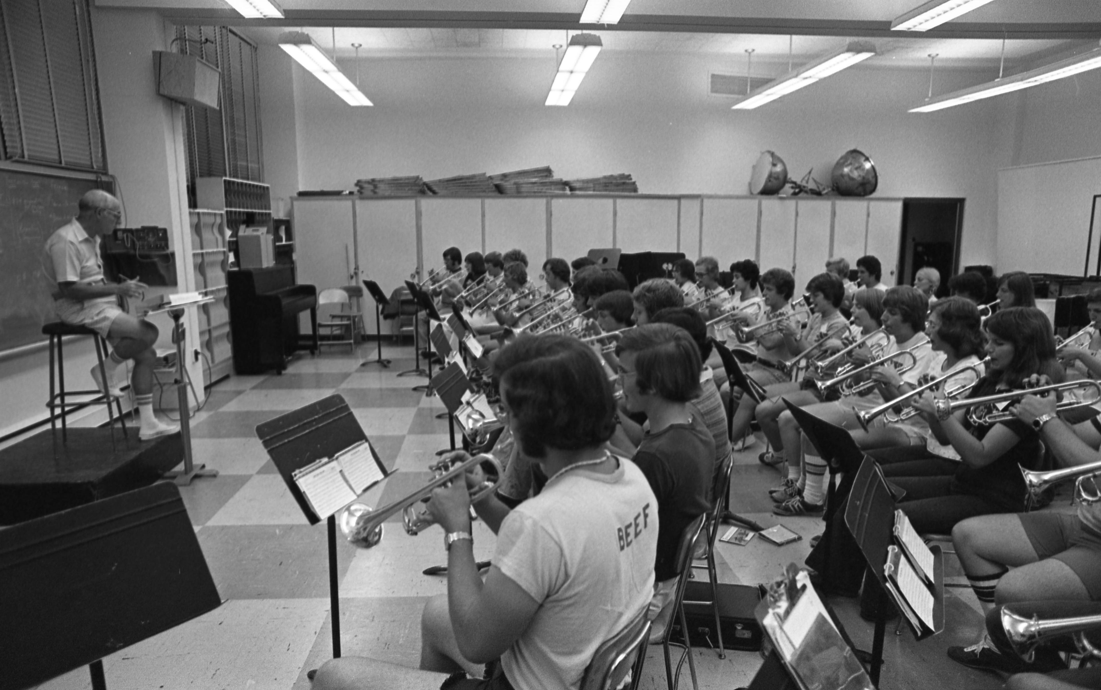 Band director Mark Kelly sits on a stool in front of a room full of trumpet players as they rehearse.