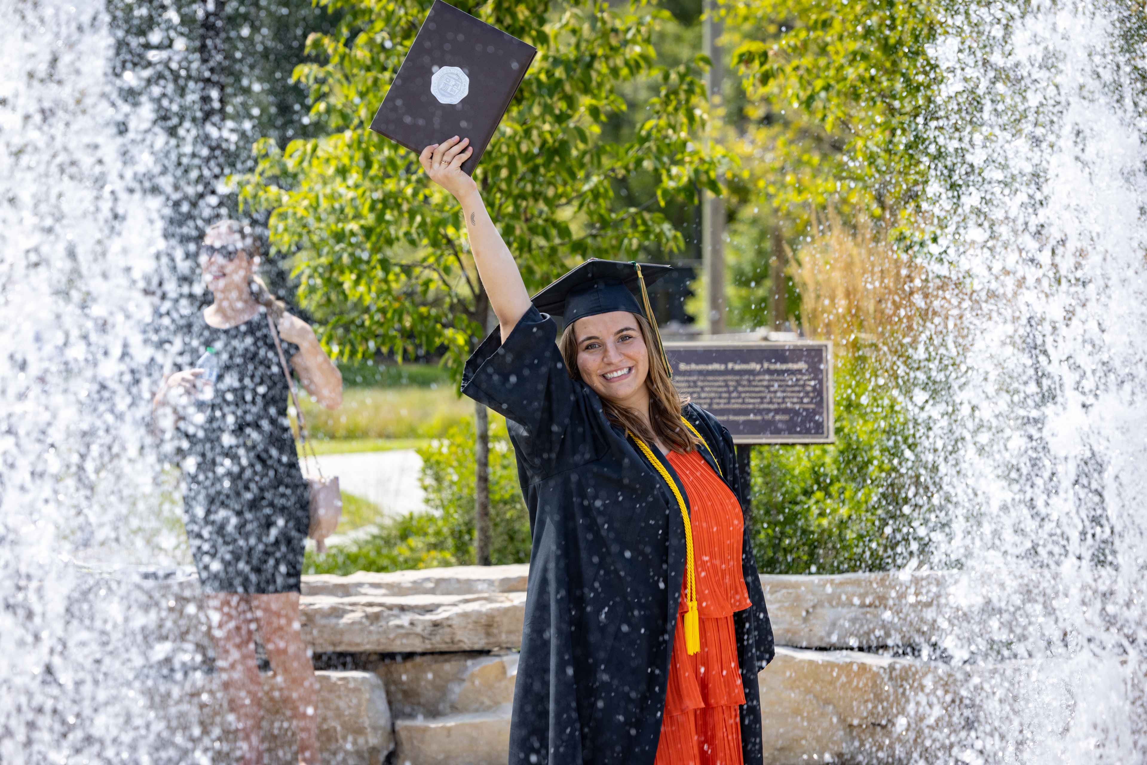 Female graduate holds diploma cover near Schmidt Family Fountain 