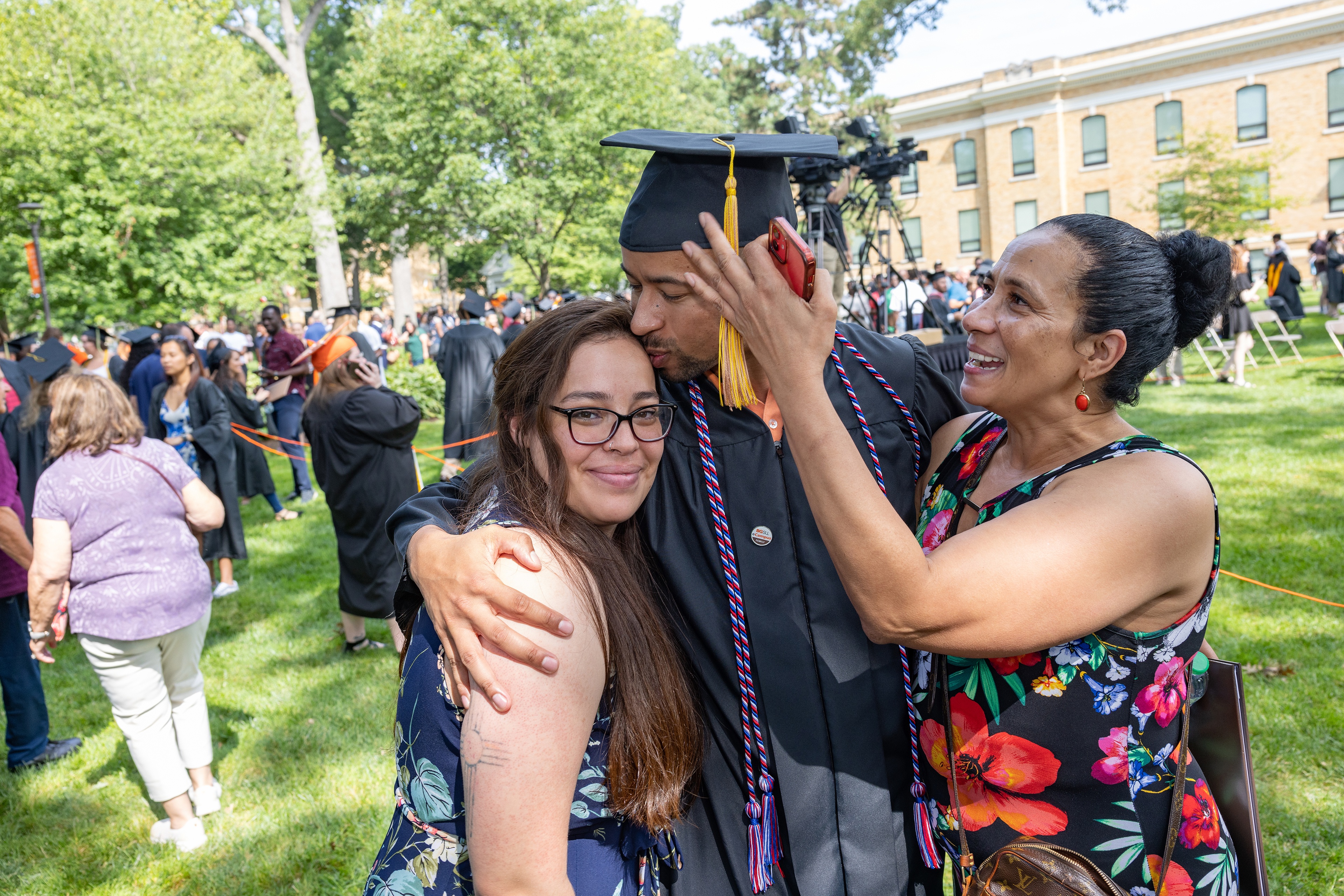 Graduate in cap and gown kisses a family member while being hugged by another person