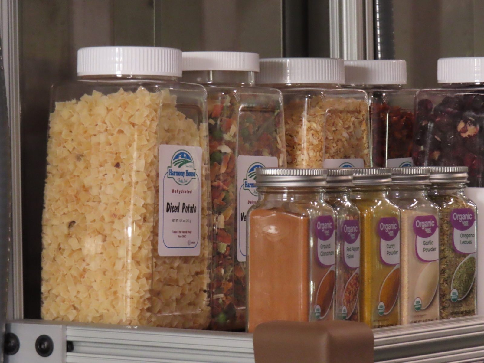 A close-up of the food storage in the kitchen of the Space Analog for the Moon and Mars. Four large jars of dehydrated food including diced potato are visible. Five small jars of spices cinnamon, red pepper flakes, curry powder, garlic powder and oregano leaves are neatly lined up on a shelf in front of the large jars. 