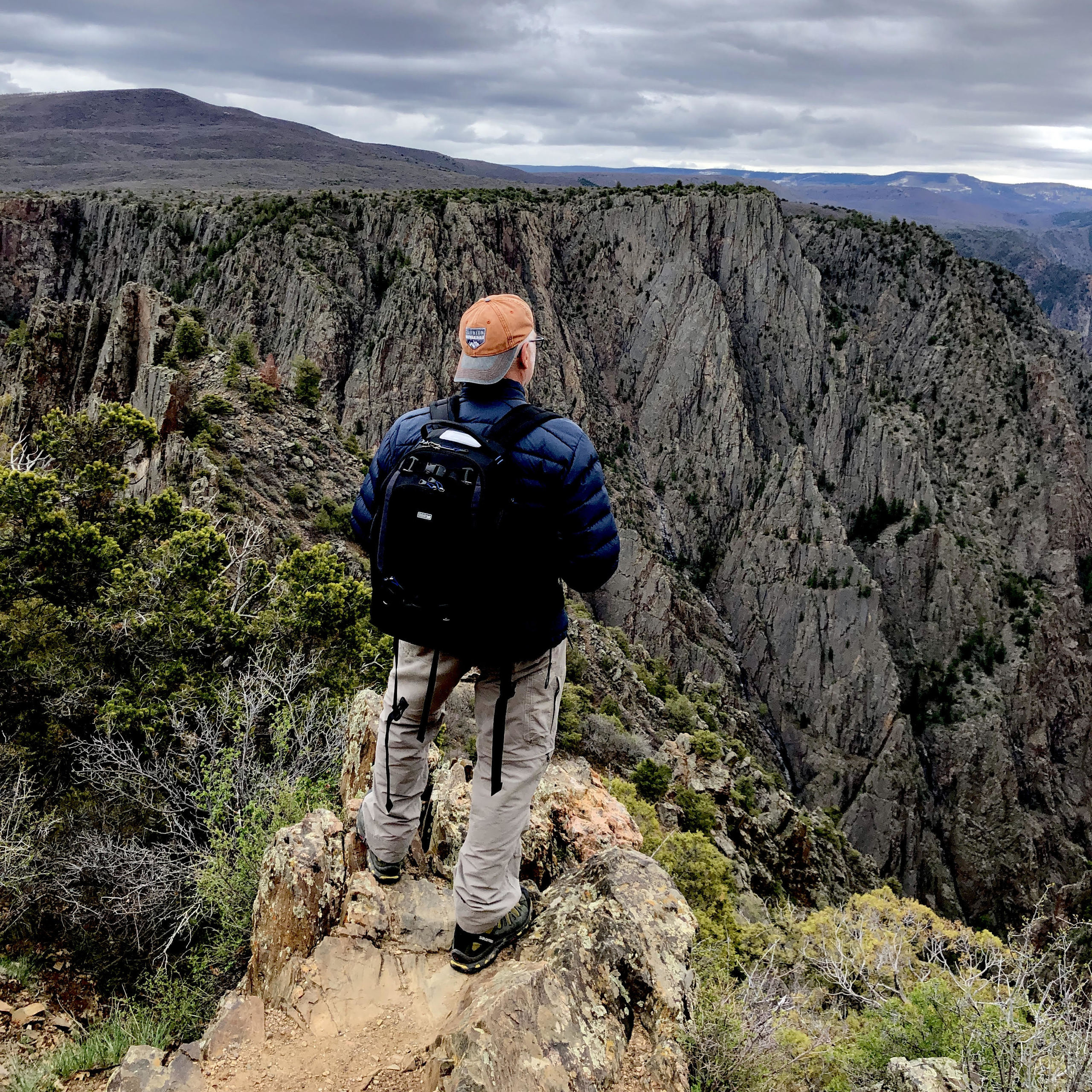 A man stands at the top of a mountain and looks over the sweeping mountain vista. His back is to the camera. 