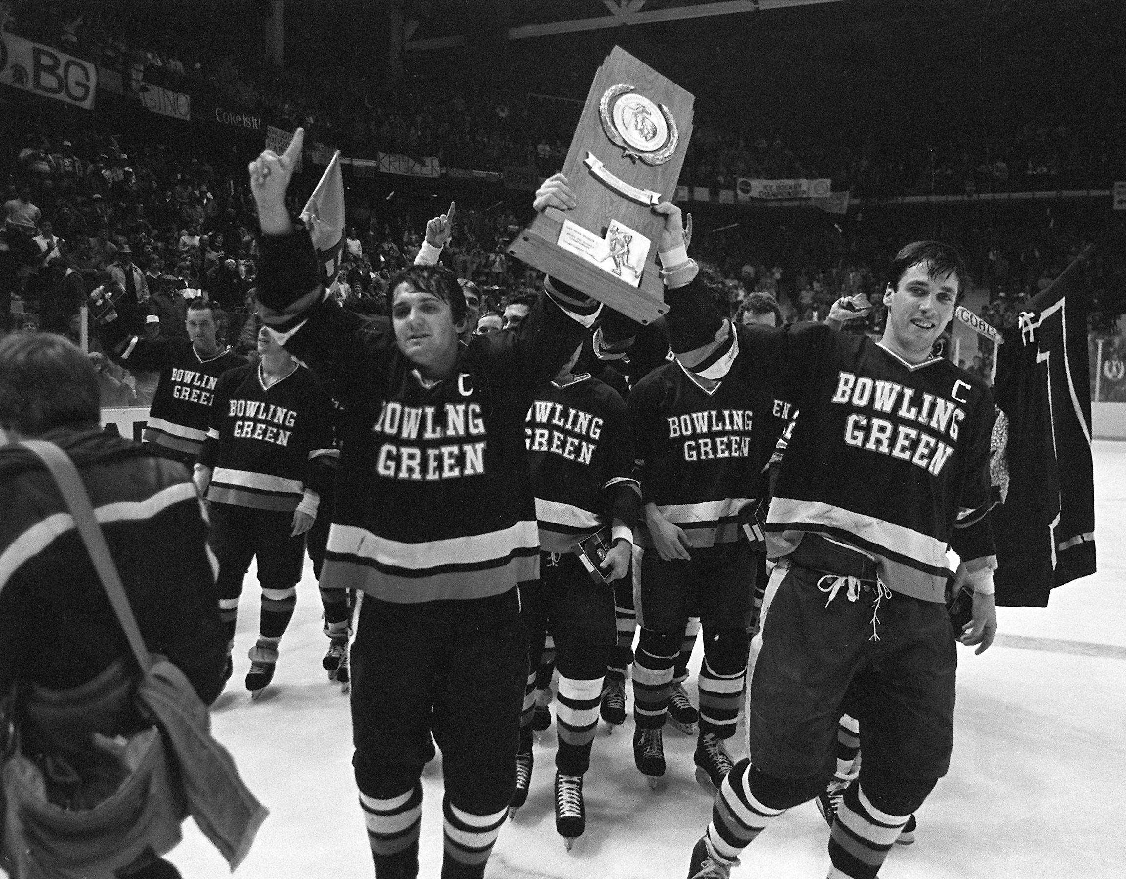 Two hockey players, Mike Pikul and Wayne Wilson, both hold an NCAA championship trophy in a black-and-white photo from 1984.