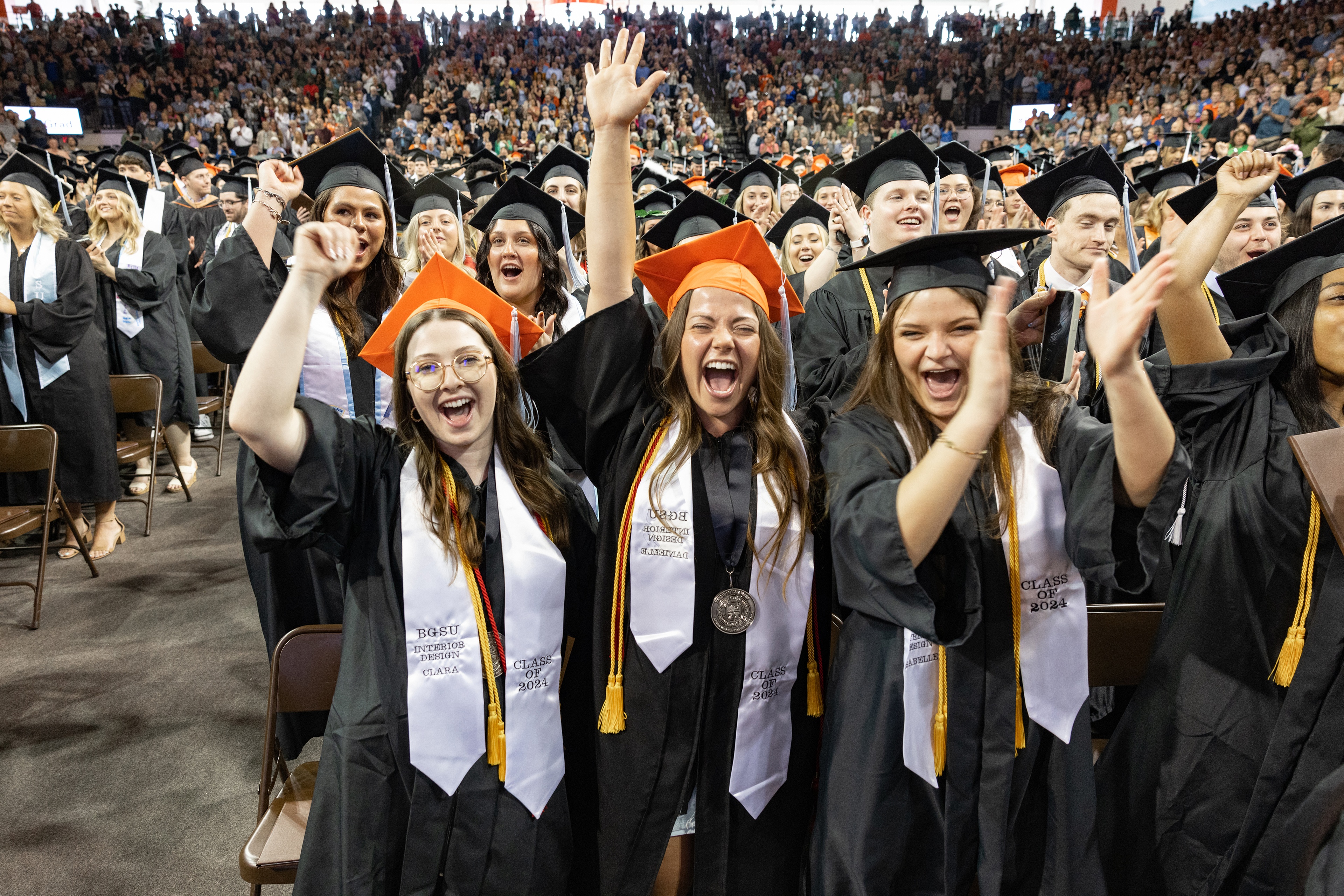 Three female graduates cheer among a crowd of graduates