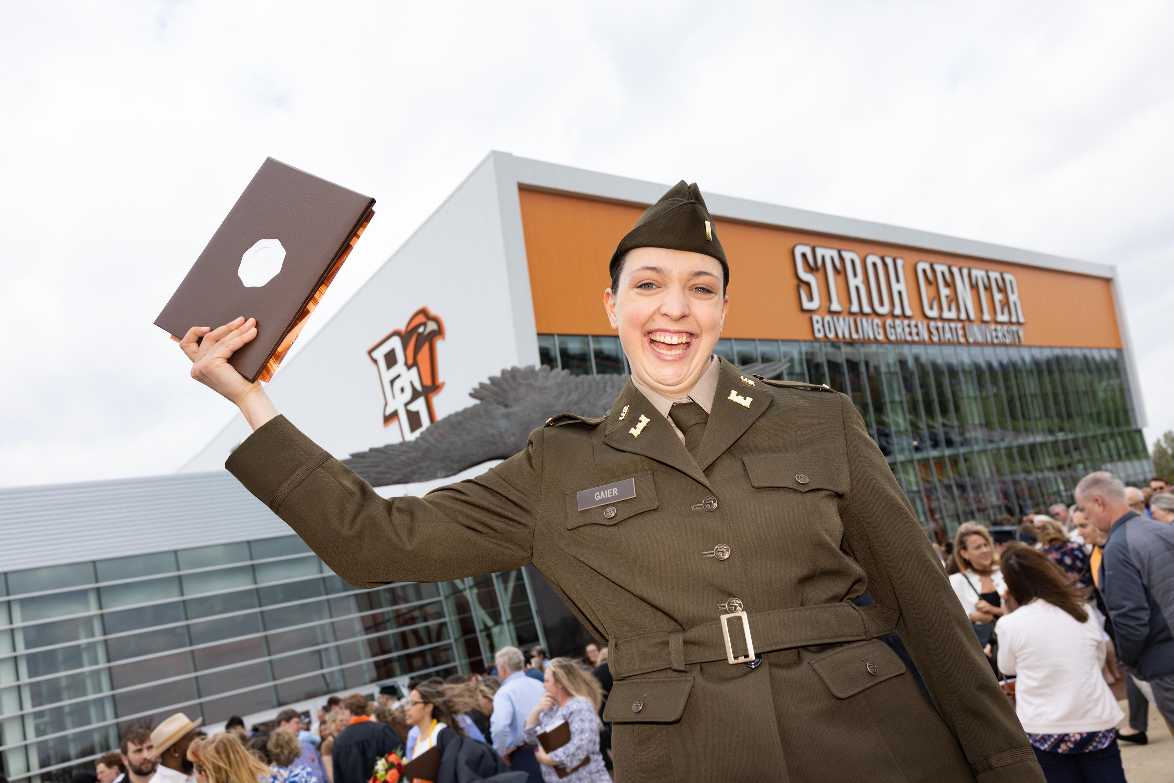 Young woman in an Army uniform holds her diploma cover high outside the Stroh Center