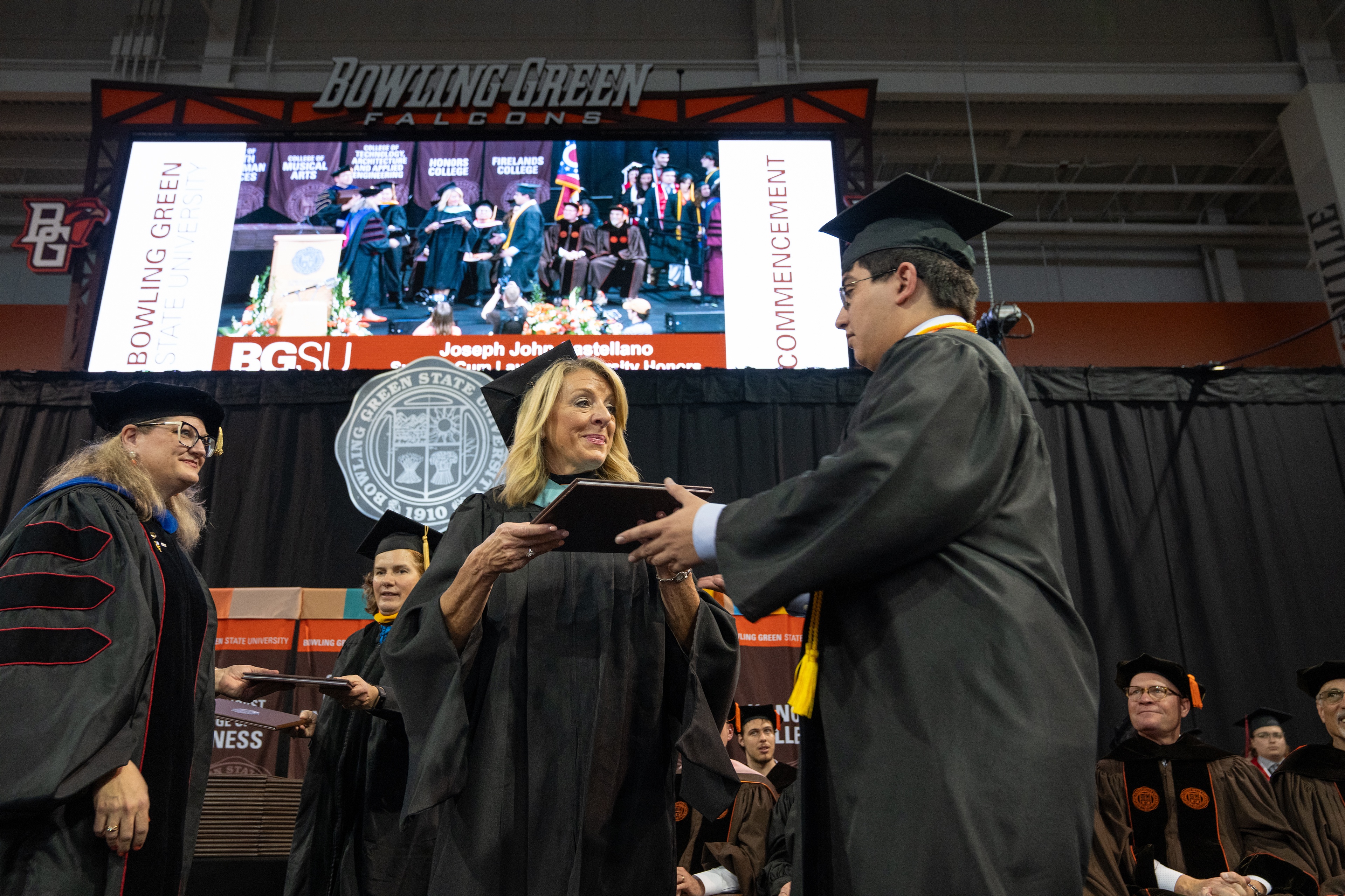 A woman in graduation regalia presents a diploma cover to a male student 