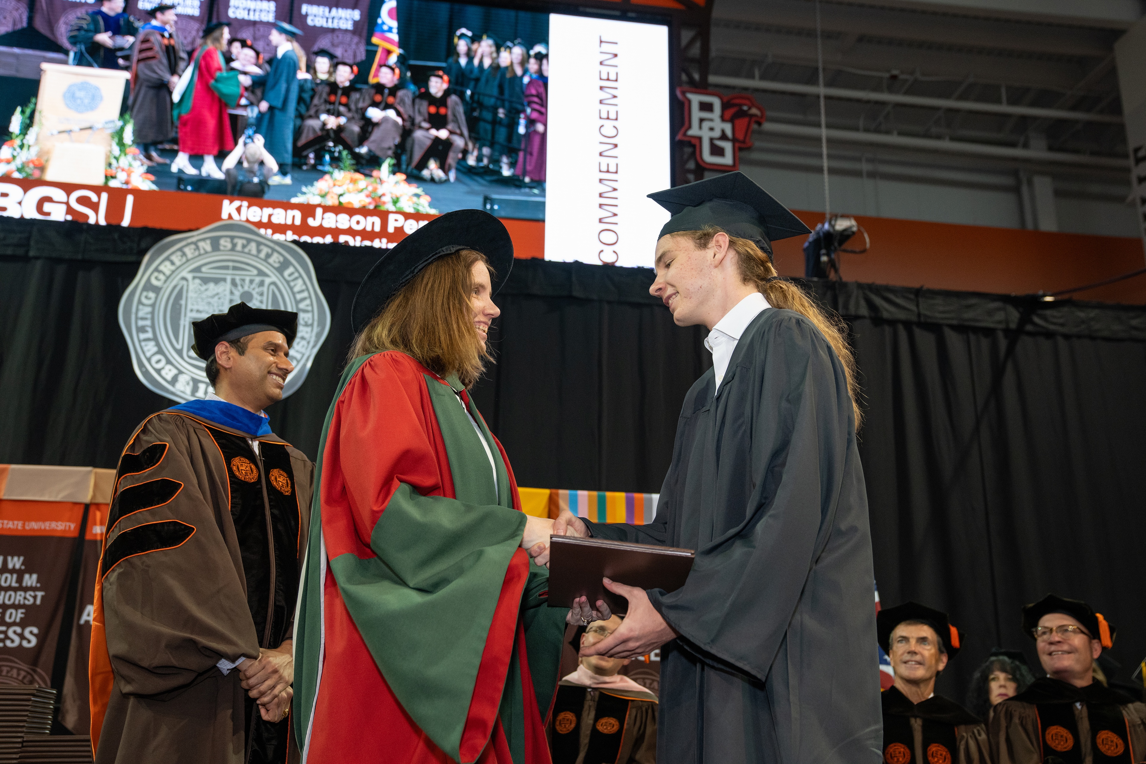 A woman in graduation regalia presents a diploma cover to a male student 