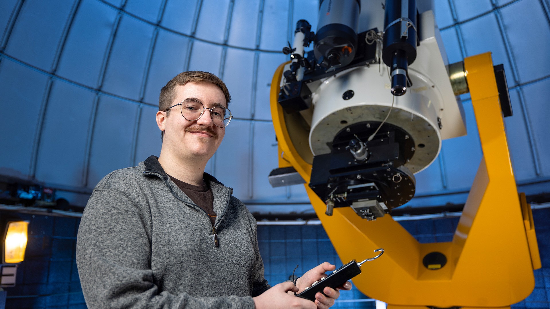 A student with a remote smiles in front of a telescope.