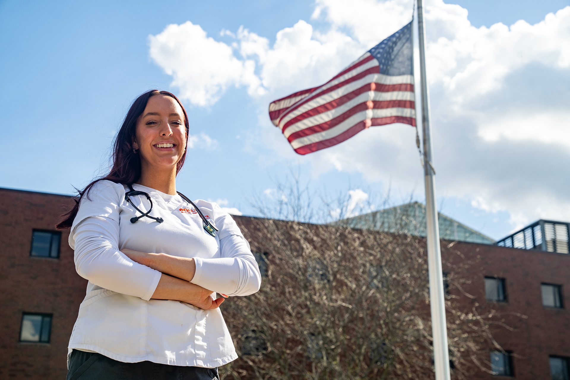 A woman poses for a picture outside with a stethoscope on and an American flag in the background.