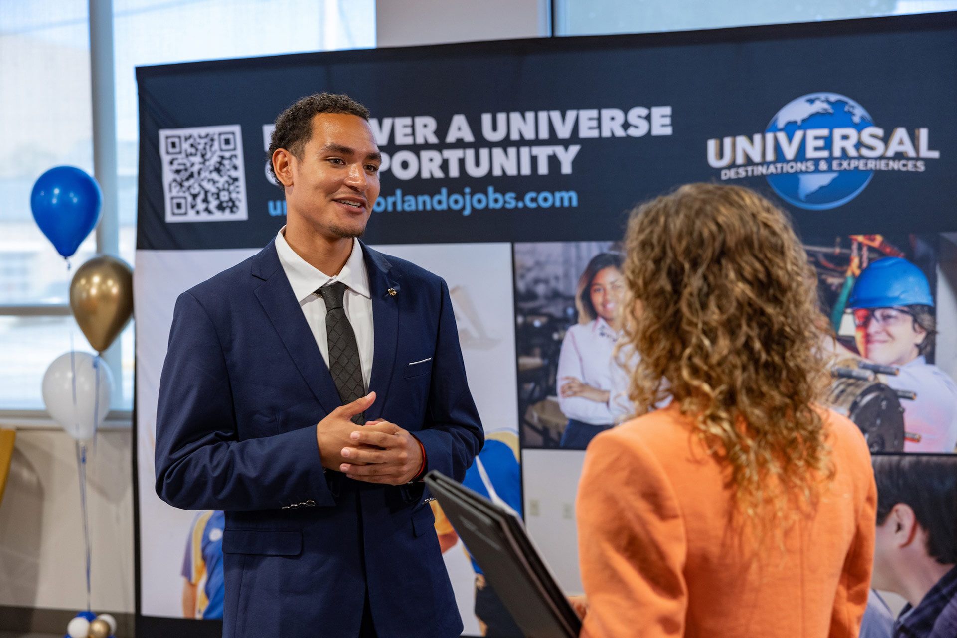 A person wearing a suit speaks to another person in front of a Universal Orlando banner.