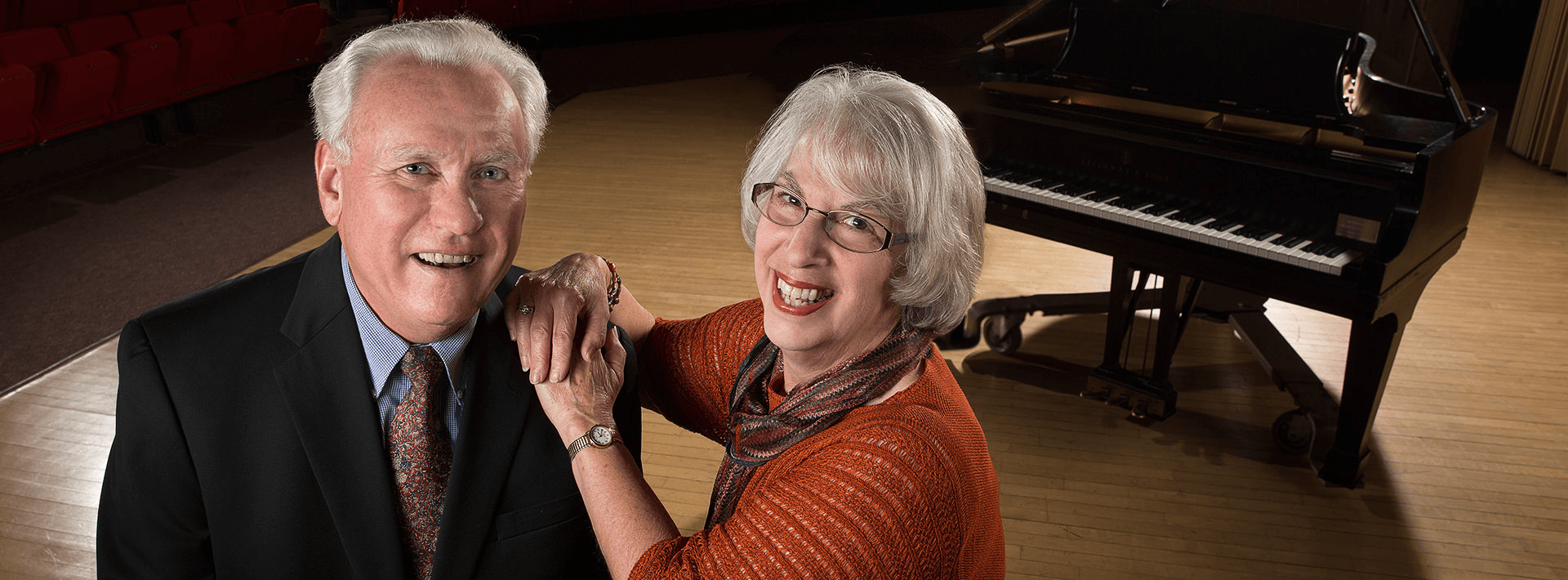 Two people pose in front of a piano.