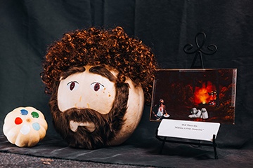 A pumpkin decorated as Bob Ross. A smaller pumpkin on the side decorated as a palette. 