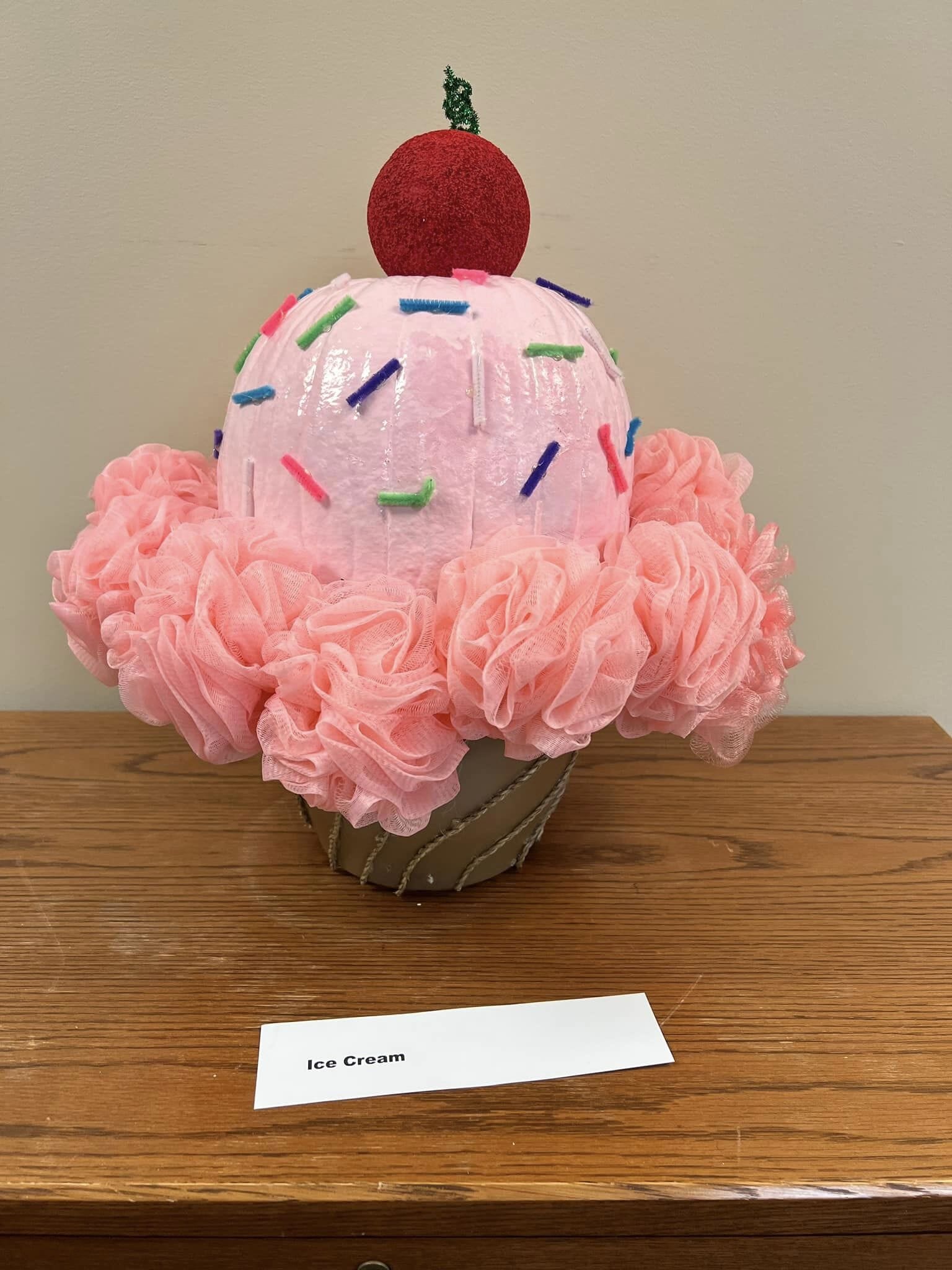A pumpkin painted and decorated as a cone with strawberry ice cream, sprinkles, and a cherry. 